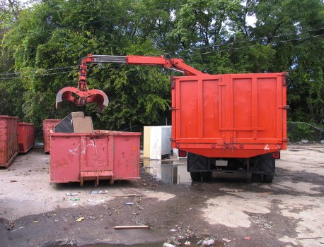A red waste collection truck equipped with a hydraulic arm and a large claw attachment is seen lifting a metal waste bin filled with rubbish. The truck is positioned on a paved surface with some scattered debris visible on the ground. To the left, there is a smaller, open-top bin also filled with waste, showing signs of wear with faded paint and scratches. In the background, dense green trees provide a natural backdrop, suggesting the scene is outdoors, possibly near a commercial or industrial area in Aldgate. The environment appears to be a typical waste management site with multiple bins and the tools used for rubbish collection, reflecting the services offered by Commercial Waste Aldgate within the local area of the postcode. The lighting indicates daytime with natural sunlight illuminating the scene clearly, highlighting the textures of the metal containers and the truck's paintwork, all consistent with a professional rubbish removal operation.