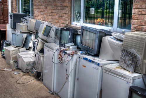 A row of old, discarded computer monitors and CRT televisions is lined up outside a brick building, likely for rubbish collection or disposal. The screens vary in size, with some being large cathode ray tube models, and others smaller. Many of the units have visible cables and wires hanging loosely or disconnected, indicating they are no longer in use. The devices display different shades of grey and black, with some showing minor signs of wear, such as dust or scratches. They are placed directly on the ground, which appears to be a concrete or paved surface, adjacent to a brick wall and beneath a large window reflecting nearby greenery. To the right, a small grey portable fan rests on top of one of the appliances. The overall scene suggests an outdoor area designated for clearing electronic waste as part of rubbish removal services, and it reflects the type of waste collected by companies like Commercial Waste Aldgate in the London area, close to the postcode district CC1 or nearby W1. The lighting is natural, indicating daytime, and the setup appears to be awaiting collection or recycling within the context of commercial waste management.