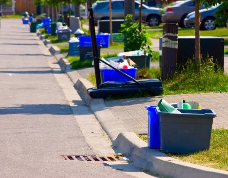 A grey plastic bin with a green lid, partially open, placed on a grass verge next to a paved sidewalk in an urban residential area. Behind the bin is a blue recycling bin and a black bin, with various discarded items visible inside. Further along the pavement, multiple blue and black rubbish bins and recycling containers are lined up on the sidewalk adjacent to parked cars and small green trees, indicating regular waste collection. The scene is illuminated by natural daylight, and the bins are positioned along a typical street in a town or city, consistent with waste management services provided by Commercial Waste Aldgate in the postcode area, possibly in London. The pavement is clean, and the environment appears well-maintained, with some greenery and grass verge separating the bins from the thoroughfare, highlighting typical rubbish disposal arrangements outside residential properties or commercial premises in the vicinity of Aldgate.