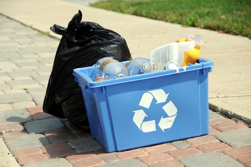 On a paved sidewalk near a grassy verge, there is a bright blue plastic recycling bin with a white recycling symbol on its side, filled with crumpled plastic bottles and containers, some with visible labels. Next to the bin, a black plastic rubbish bag is placed and appears to be partially filled, tied at the top. The scene suggests a collection point for waste disposal or recycling, likely outdoors in a residential or commercial area within or near Aldgate, London, as part of a rubbish removal service by Commercial Waste Aldgate. The background features a well-maintained sidewalk and green grass which contributes to a tidy environment. The lighting indicates natural daylight, emphasizing the clean, orderly presentation of waste materials prepared for collection or disposal in accordance with local waste management practices.