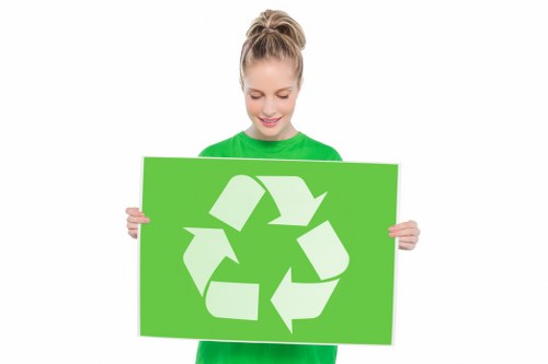 A young woman with blonde hair tied in a high ponytail, dressed in a bright green T-shirt, standing indoors against a plain white background, holding a large, rectangular green sign with a white recycling symbol comprising three arrows forming a triangle, symbolising recycling and waste management. Although the focus is on her smiling face and the sign, the image emphasizes environmental awareness and responsible waste disposal. The setting is minimalistic, with no visible environmental or waste-related objects, but the recycling symbol connects to rubbish removal services offered by companies like Commercial Waste Aldgate, serving areas around London and postal code E1. The overall tone is neutral and professional, suited for conveying eco-friendly waste management practices online.