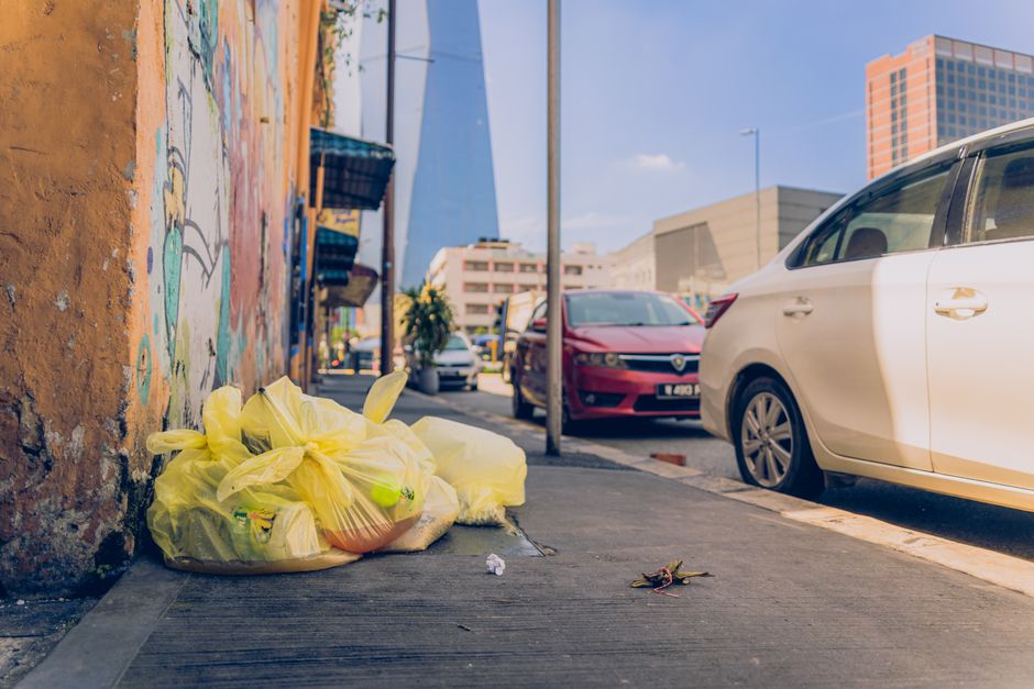 A collection of overflowing waste bins and piles of rubbish positioned on a paved urban street corner under natural daylight. The scene includes a large grey bin with a partially open blue lid, spilli