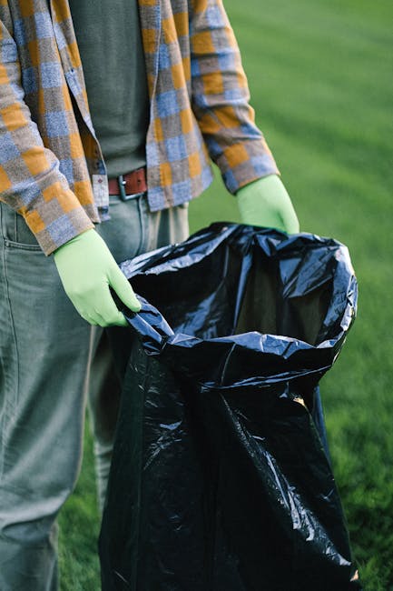 A waste collection worker wearing a high-visibility vest and dark trousers is operating a large red refuse collection vehicle parked on the side of a street. The worker is standing behind the open rea