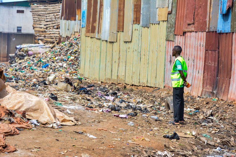 A sanitation worker in a yellow and red high-visibility jacket is operating a large red rubbish collection vehicle parked on the side of a street. The vehicle's rear hopper, used for waste compaction,